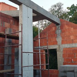 TP - BUD TOMASZ PIĄTEK - Wznoszenie ścian z czerwonej cegły na budowie domu, widoczne podpory i betonowe elementy konstrukcyjne. W tle zieleń drzew.