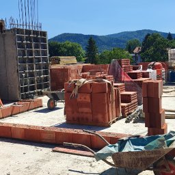 TP - BUD TOMASZ PIĄTEK - Budowa domu z czerwonej cegły: wznoszone ściany, szalunek żelbetowy, palety cegieł, taczki. W tle zieleń i zabudowa mieszkalna w słoneczny dzień.