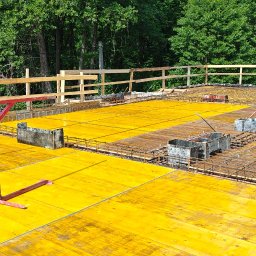 TP - BUD TOMASZ PIĄTEK - Żółte szalunki i zbrojenie na budowie, z pracującym zbrojarzem w tle, otoczone zielenią drzew. Widoczne metalowe pojemniki i czerwone podpory szalunkowe.
