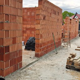 TP - BUD TOMASZ PIĄTEK - Wznoszone ściany z czerwonej ceramiki poryzowanej na fundamencie z izolacją. Widoczne narzędzia i materiały budowlane. Pochmurne niebo w tle.