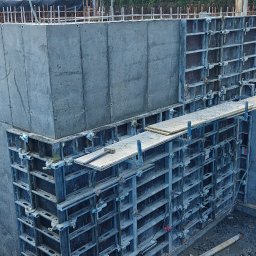 TP - BUD TOMASZ PIĄTEK - Szare, betonowe ściany fundamentowe z widocznymi otworami okiennymi i elementami szalunków, częściowo zbrojone, na placu budowy w pochmurny dzień.