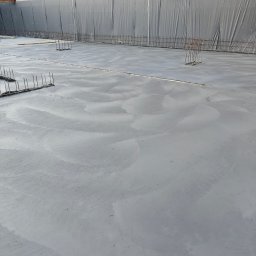 TP - BUD TOMASZ PIĄTEK - Wylany fundament z wystającymi prętami zbrojeniowymi, zabezpieczony folią od strony gruntu. Widoczne ślady po zacieraniu betonu.