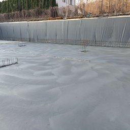 TP - BUD TOMASZ PIĄTEK - Świeżo wylany fundament z wystającymi prętami zbrojeniowymi, zabezpieczony folią od strony skarpy. Widoczne ślady po rozprowadzaniu betonu.