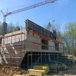 TP - BUD TOMASZ PIĄTEK - Surowy budynek żelbetowy w trakcie budowy, otoczony rusztowaniami i dźwigiem, na tle zieleni i błękitnego nieba. Widoczne szalunki i materiały budowlane.