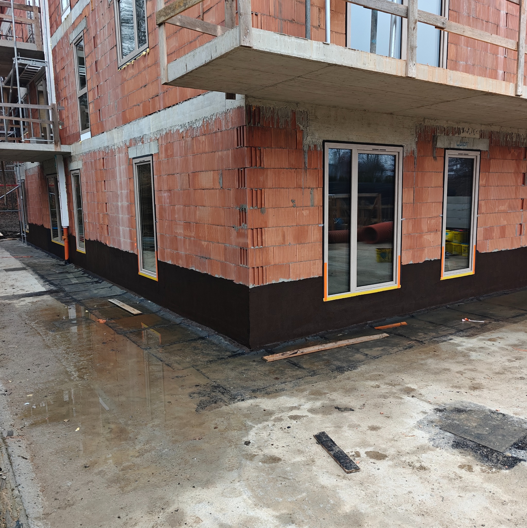 Ściana budynku z czerwonej cegły z zaizolowanymi fundamentami, widoczne okna z żółtą taśmą zabezpieczającą. Na ziemi woda, w tle rusztowanie.