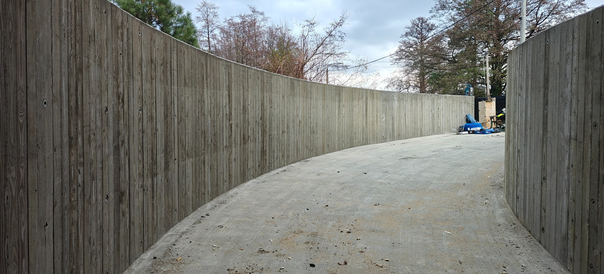 Zakrzywiona ściana z betonu architektonicznego o fakturze drewna, widoczna z perspektywy chodnika. W tle drzewa i niebo.