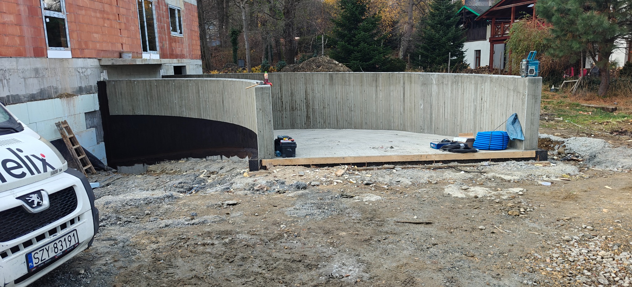 Żelbetowa konstrukcja garażu podziemnego w trakcie budowy, widoczna izolacja, narzędzia i materiały budowlane. W tle budynek w stanie surowym otwartym i zieleń.