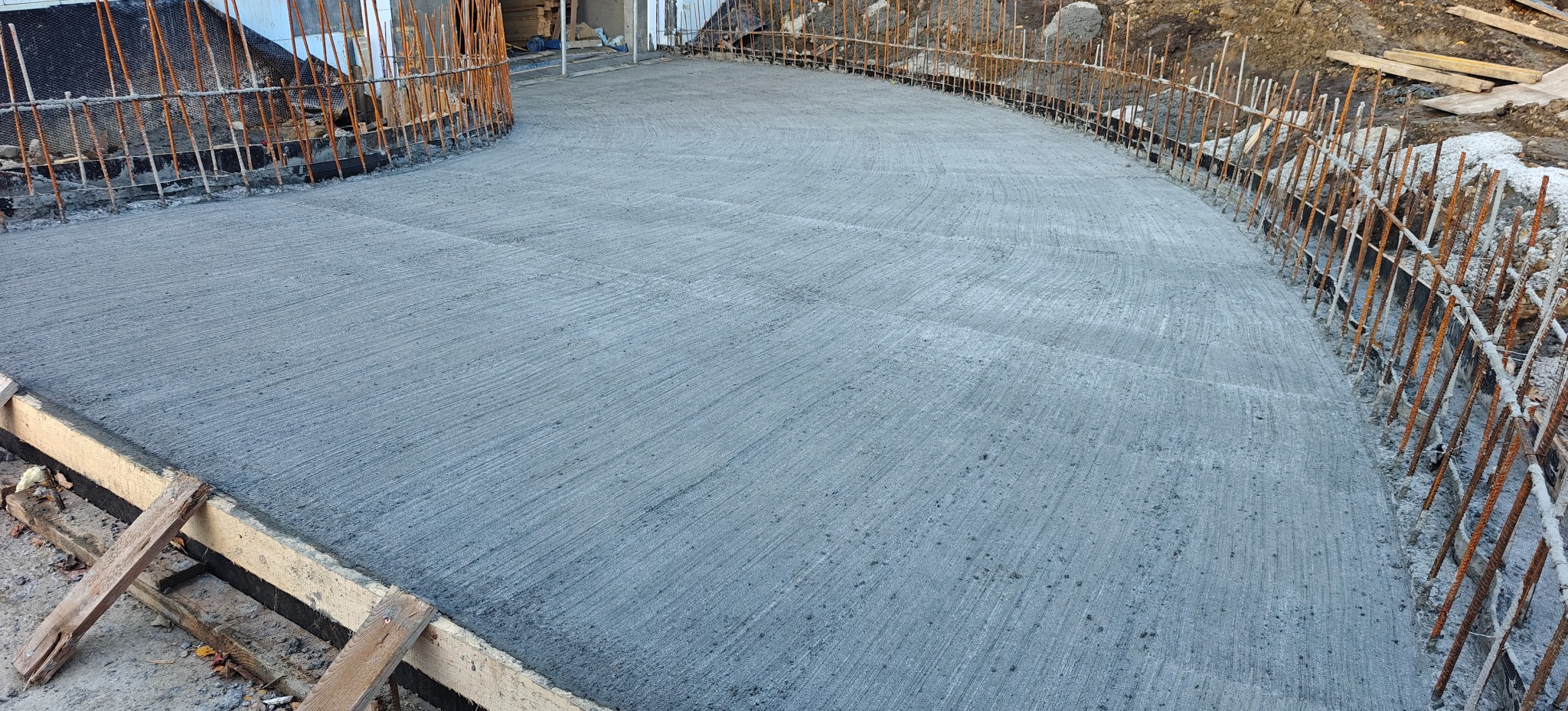 Świeżo wylany, gładki betonowy fundament z widocznym zbrojeniem po bokach, przygotowany do dalszych prac budowlanych. Widok z góry, oświetlenie dzienne.