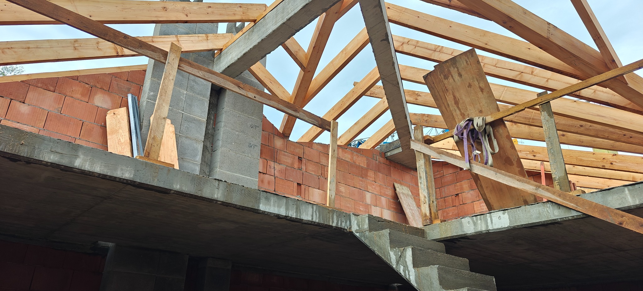 Surowy stan budynku w trakcie budowy: ściany z czerwonej cegły, betonowe schody i drewniana konstrukcja dachu pod błękitnym niebem. Widok z dołu.