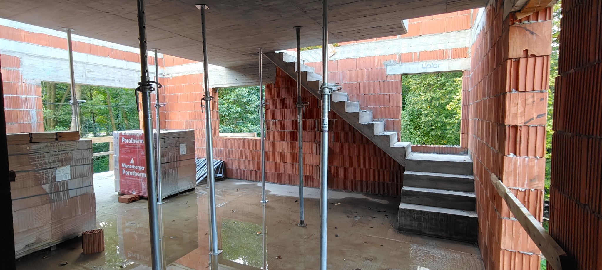 Wnętrze w trakcie budowy: murowane ściany z pustaków ceramicznych, betonowe schody, podpory stropowe, okna z widokiem na zieleń. Częściowo zalana posadzka.