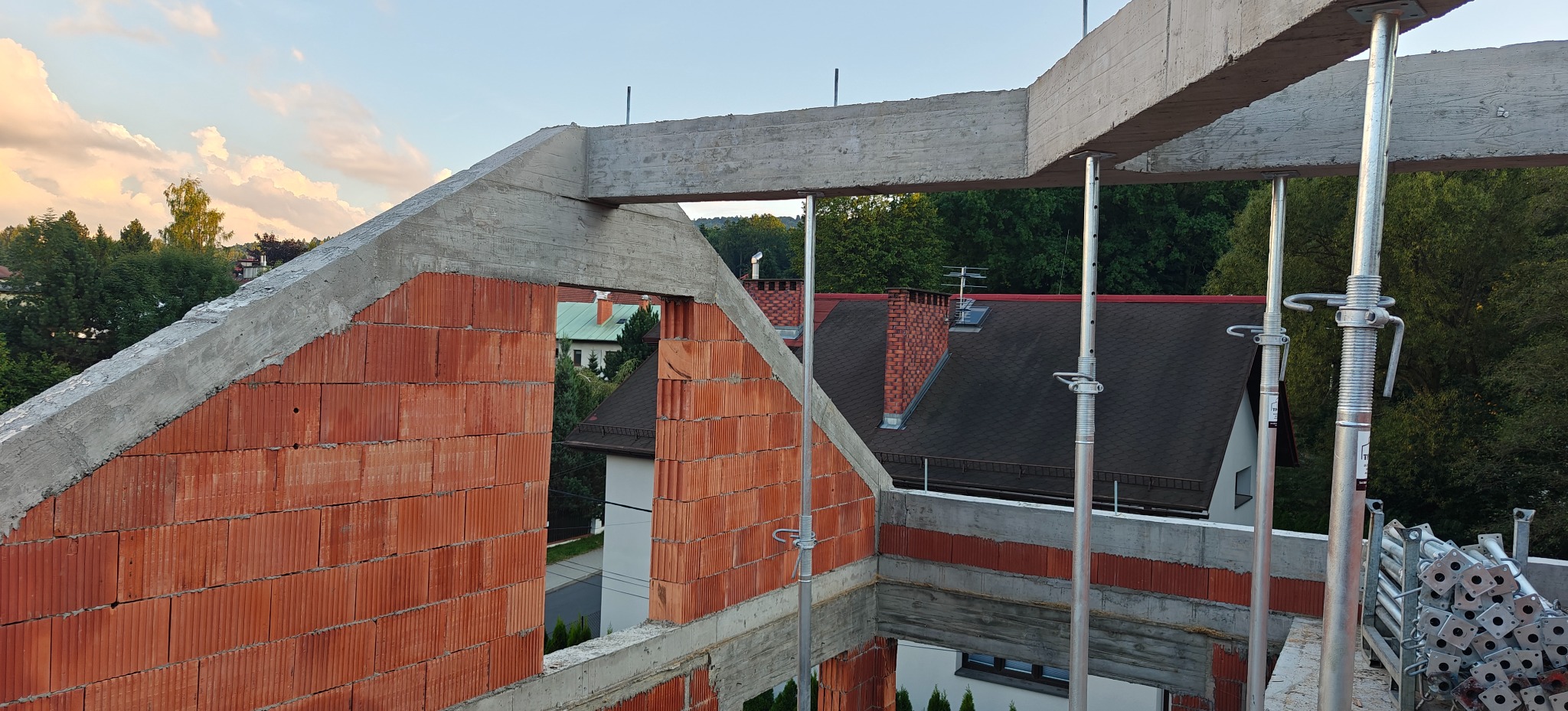 Wznoszenie ścian z czerwonej cegły na budowie domu jednorodzinnego, wsparte metalowymi stemplami. Widoczne betonowe nadproża i fragmenty konstrukcji. Tło: zabudowa mieszkalna.