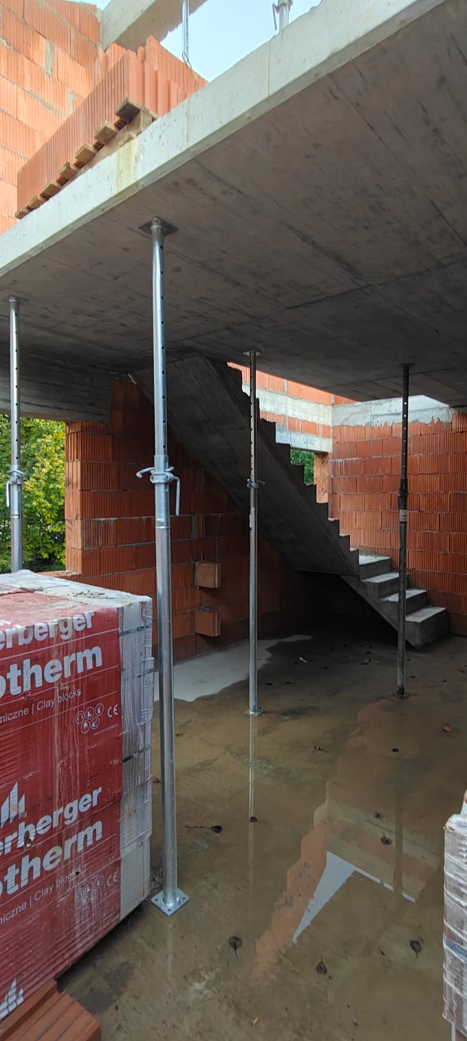 Wnętrze budynku w trakcie budowy: ściany z czerwonej cegły, betonowy strop podparty metalowymi stemplami, schody betonowe. Na pierwszym planie paleta cegieł.