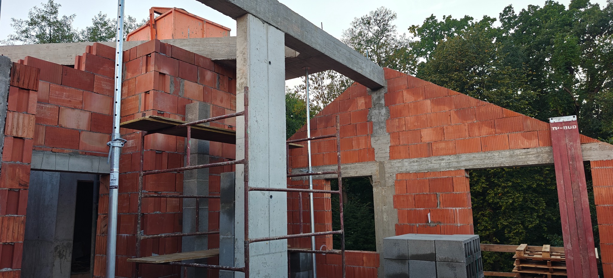 Wznoszenie ścian z czerwonej cegły na budowie domu, widoczne podpory i betonowe elementy konstrukcyjne. W tle zieleń drzew.