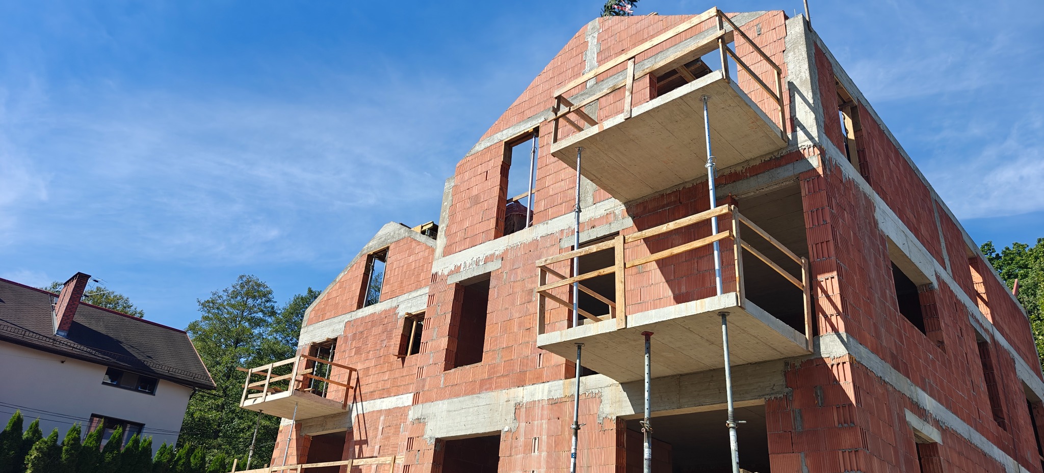Wznoszenie domu z czerwonej cegły, widoczne rusztowania i niezadaszona konstrukcja. Budynek w trakcie budowy, z otworami okiennymi i elementami betonowymi.