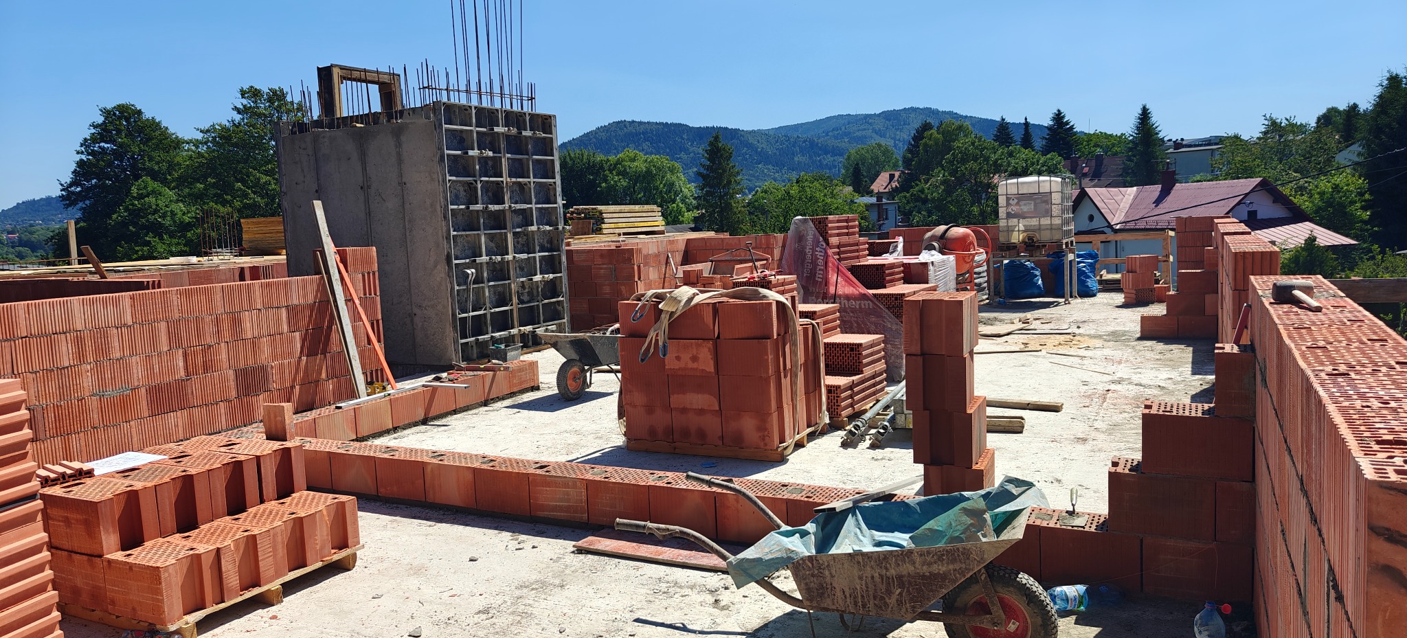 Budowa domu z czerwonej cegły: wznoszone ściany, szalunek żelbetowy, palety cegieł, taczki. W tle zieleń i zabudowa mieszkalna w słoneczny dzień.