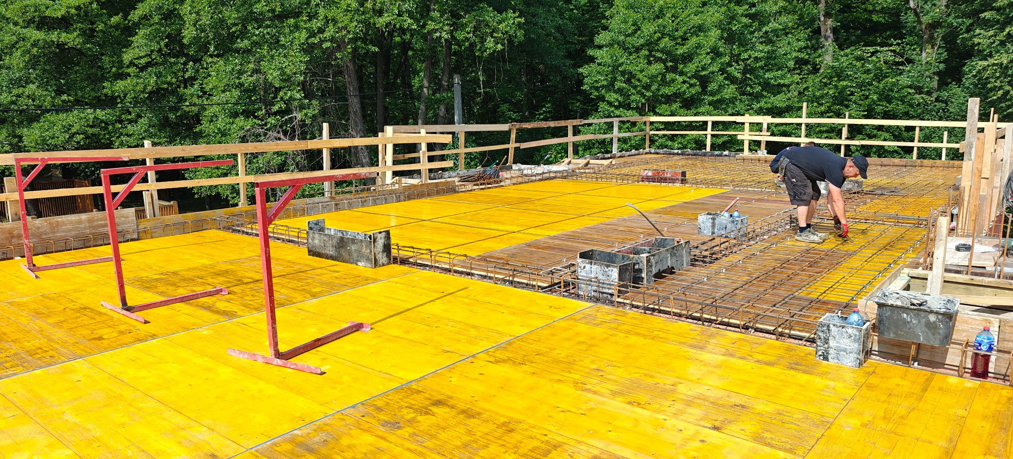 Żółte szalunki i zbrojenie na budowie, z pracującym zbrojarzem w tle, otoczone zielenią drzew. Widoczne metalowe pojemniki i czerwone podpory szalunkowe.