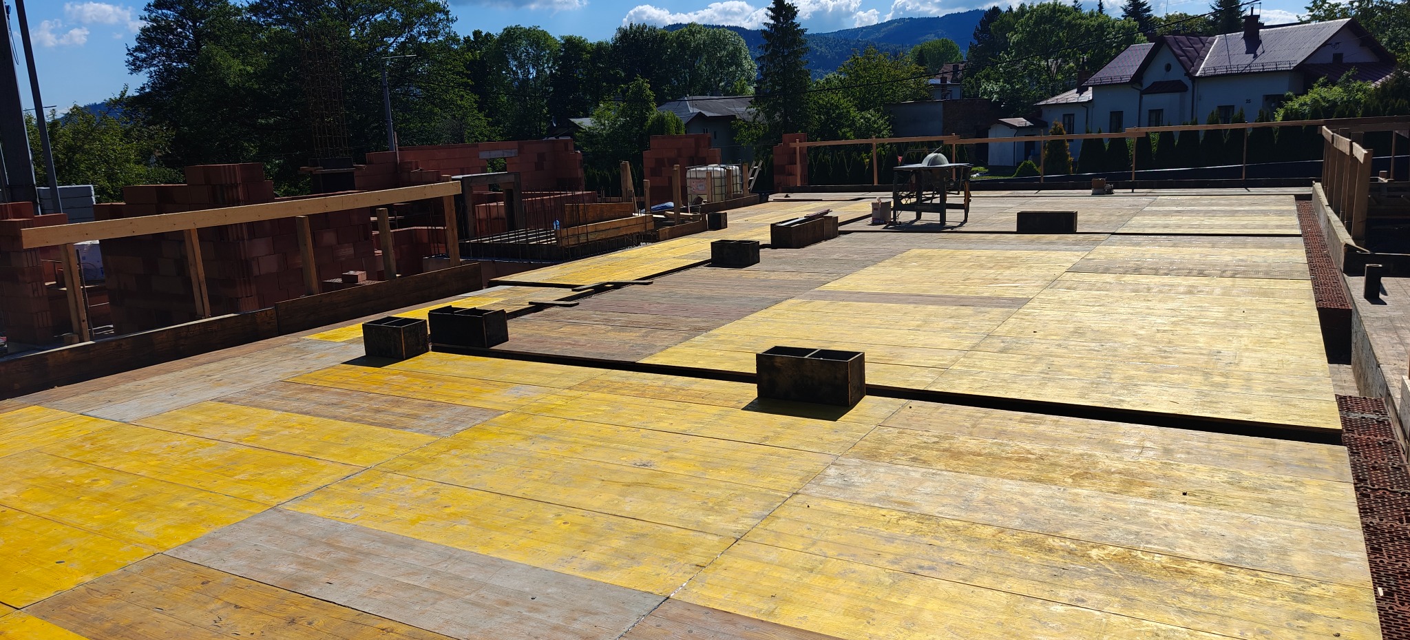 Żółte płyty szalunkowe na budowie domu z cegły, widoczne elementy konstrukcyjne i narzędzia. W tle zieleń i zabudowa mieszkalna.