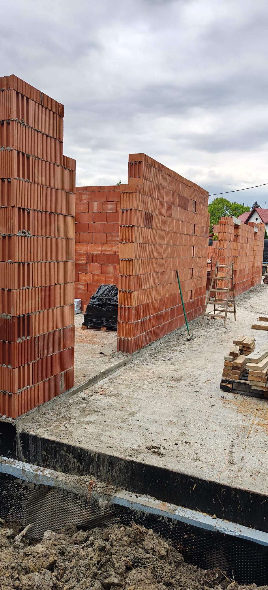 Wznoszone ściany z czerwonej ceramiki poryzowanej na fundamencie z izolacją. Widoczne narzędzia i materiały budowlane. Pochmurne niebo w tle.