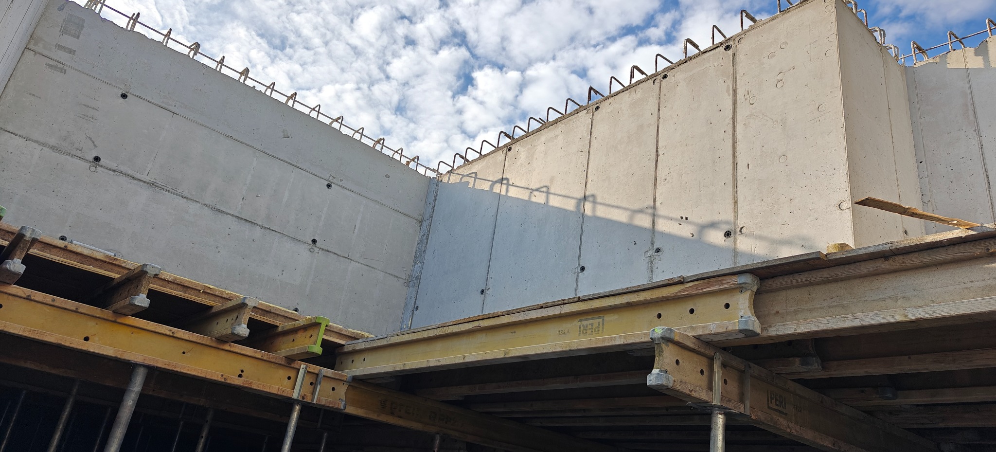 Surowe ściany żelbetowe z wystającymi prętami zbrojeniowymi, wsparte na drewnianym szalunku, na tle błękitnego nieba z chmurami. Widok z dołu.