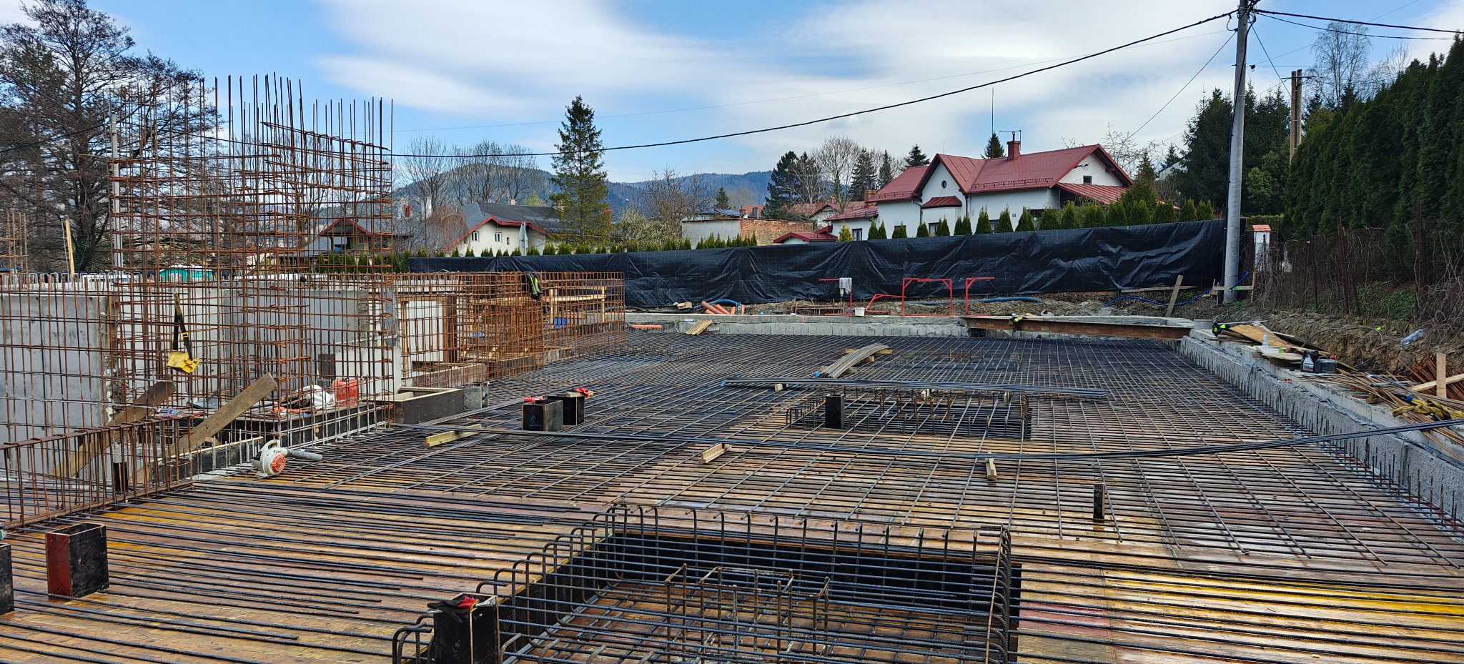 Zbrojenie fundamentów domu jednorodzinnego z widoczną siatką zbrojeniową i wystającymi prętami, przygotowane do zalania betonem. W tle zabudowa mieszkalna i błękitne niebo.