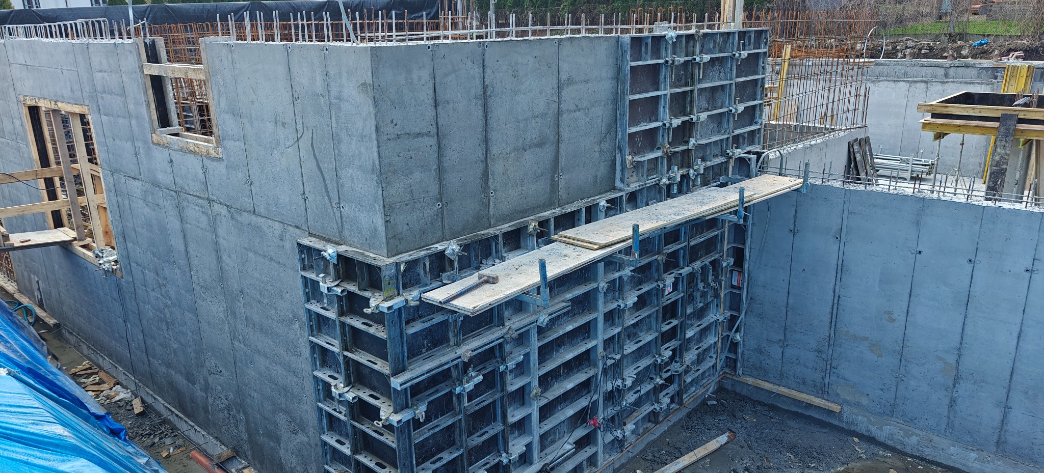 Szare, betonowe ściany fundamentowe z widocznymi otworami okiennymi i elementami szalunków, częściowo zbrojone, na placu budowy w pochmurny dzień.