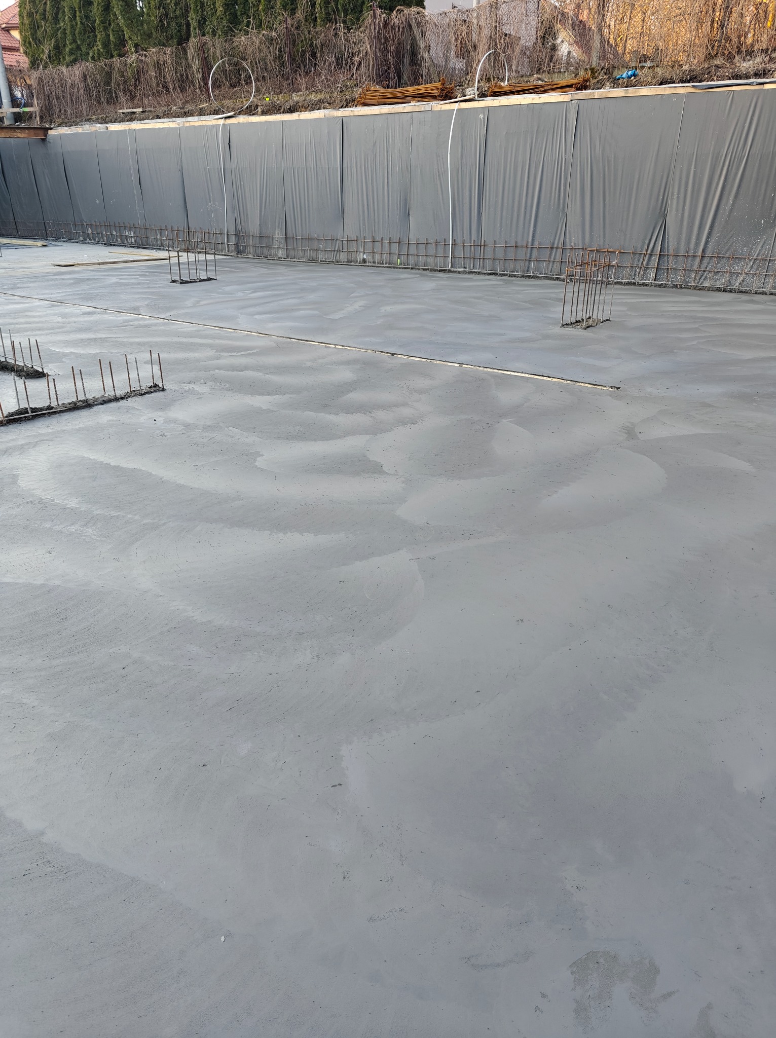 Wylany fundament z wystającymi prętami zbrojeniowymi, zabezpieczony folią od strony gruntu. Widoczne ślady po zacieraniu betonu.