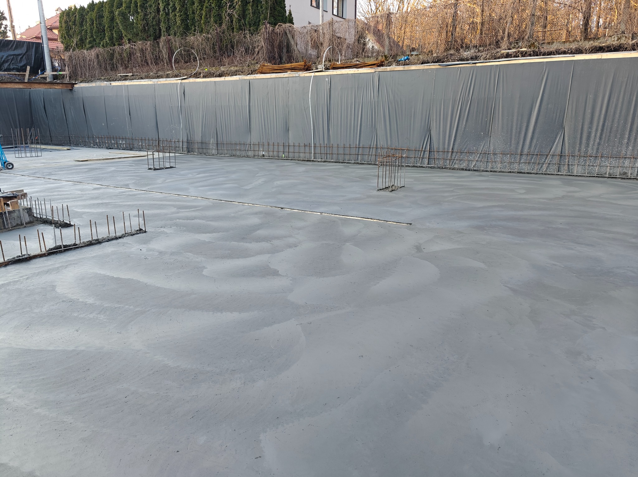 Świeżo wylany fundament z wystającymi prętami zbrojeniowymi, zabezpieczony folią od strony skarpy. Widoczne ślady po rozprowadzaniu betonu.