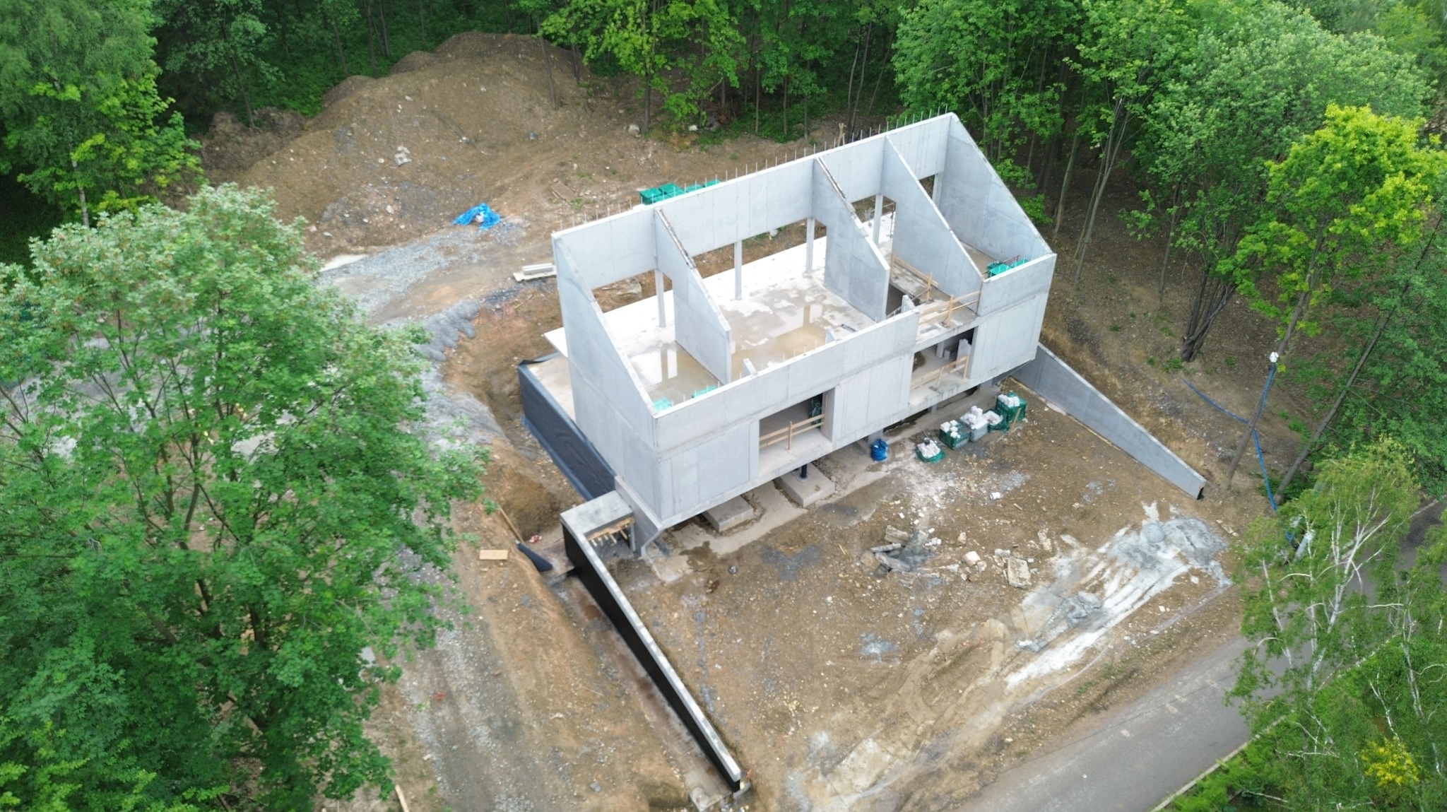Surowy, dwukondygnacyjny budynek żelbetowy w trakcie budowy, widok z góry. Widoczne fundamenty, ściany i elementy konstrukcyjne. Otoczenie: teren budowy, drzewa.