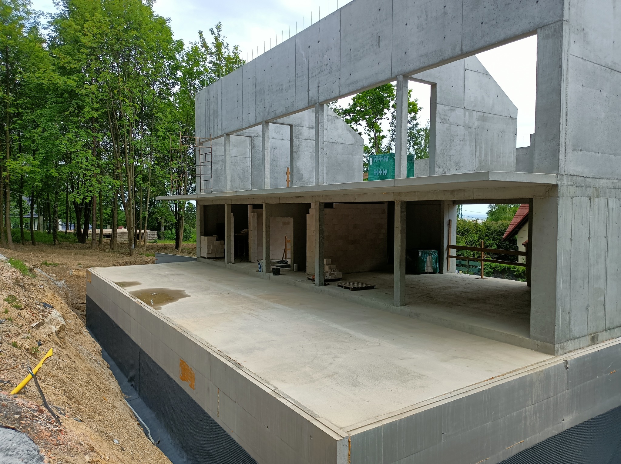 Surowy stan budynku żelbetowego w otoczeniu zieleni, widoczne fundamenty z izolacją, ściany i stropy w trakcie budowy, zarys przyszłego domu.