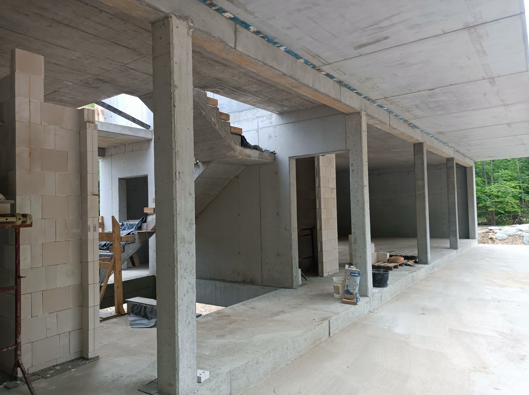Surowy stan budynku żelbetowego w Wieprzu: widoczne ściany z betonu komórkowego, betonowe słupy i strop. W tle schody w stanie surowym i zieleń.