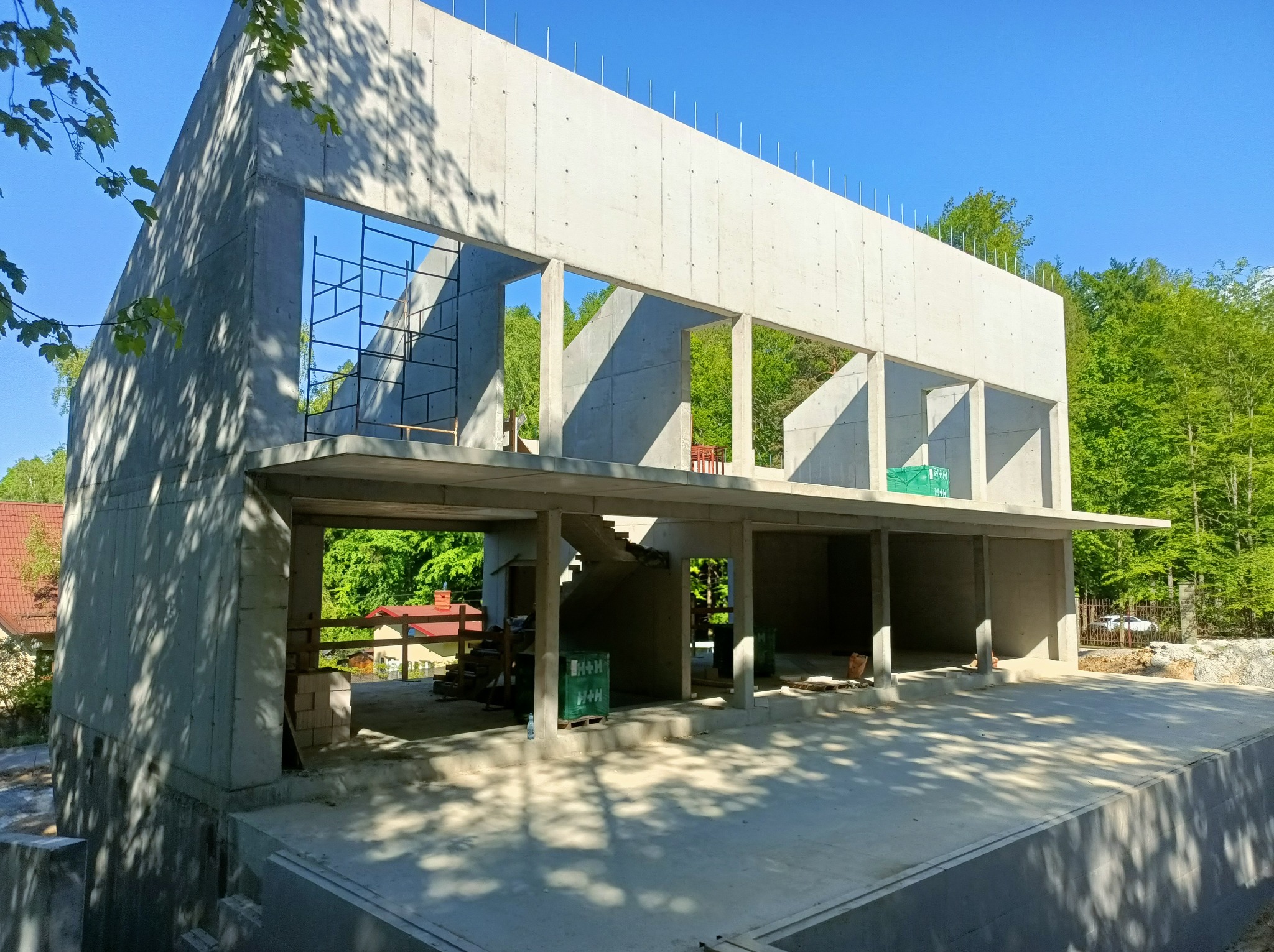 Surowy budynek żelbetowy w trakcie budowy, widoczne szalunki i zbrojenia, otoczenie zieleni, błękitne niebo. Konstrukcja w stanie nieukończonym.