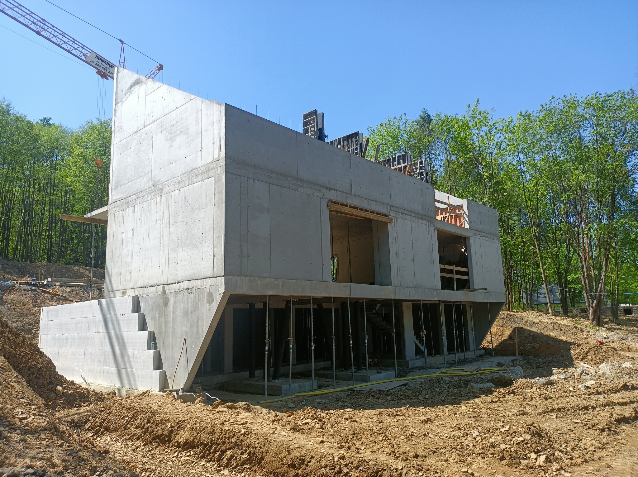 Surowy budynek żelbetowy w trakcie budowy, widoczne podpory i szalunki, zieleń w tle, jasne niebo. Ujęcie z dołu podkreśla masywność konstrukcji.