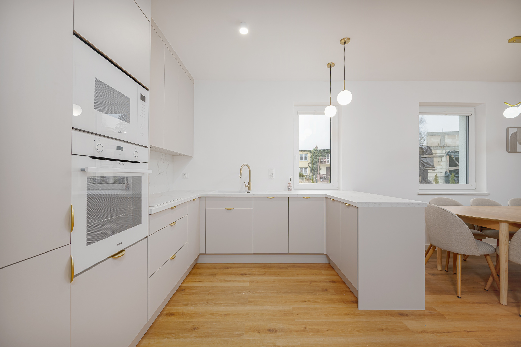 Jasna kuchnia w stylu minimalistycznym z drewnianą podłogą, złotymi uchwytami i lampami. Widoczne okna z widokiem na budynek. Sprzęt AGD marki Amica.
