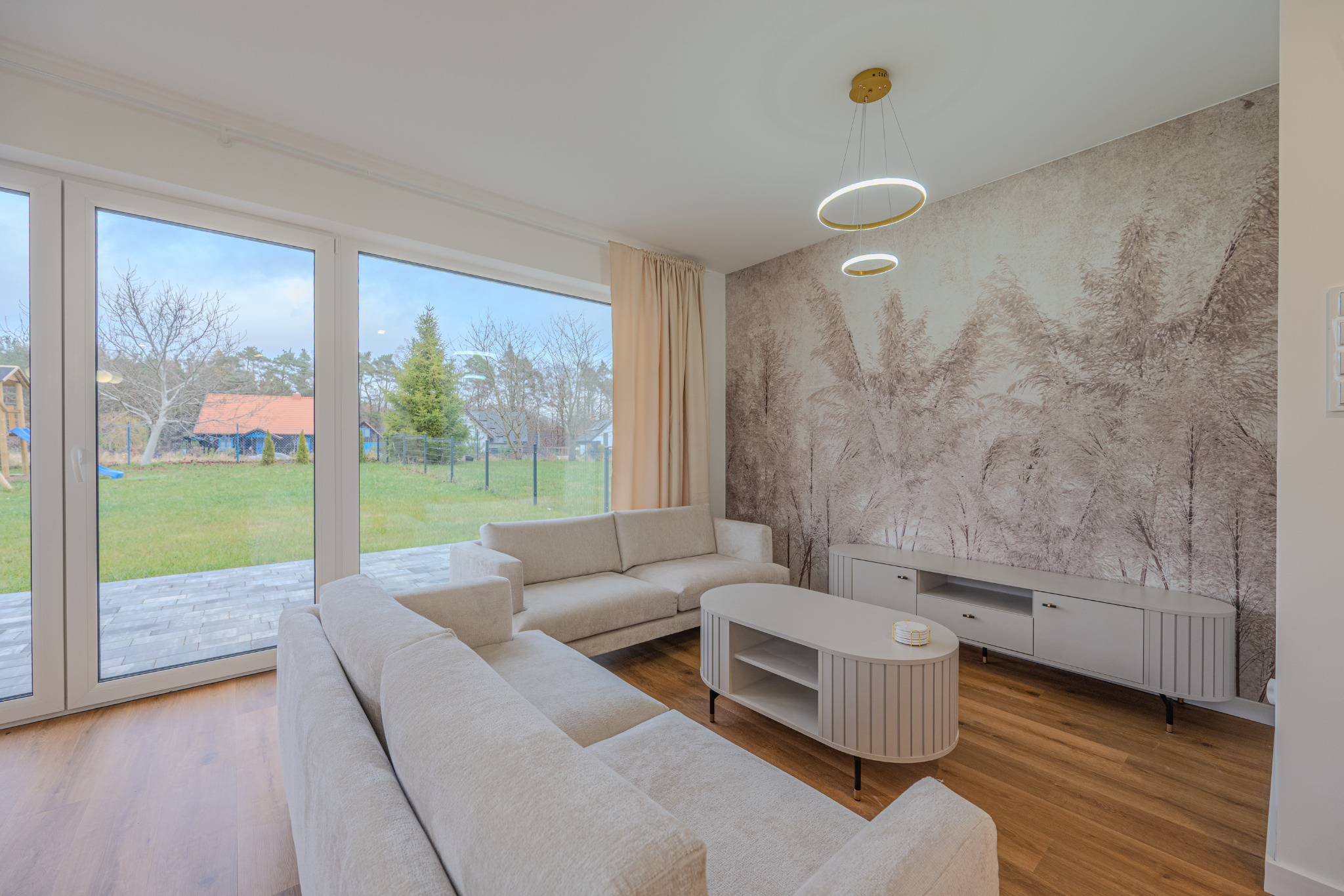 Jasny salon z beżową sofą narożną, owalnym stolikiem i szafką RTV na tle ściany z motywem roślinnym. Duże okno z widokiem na ogród. Nowoczesne oświetlenie.