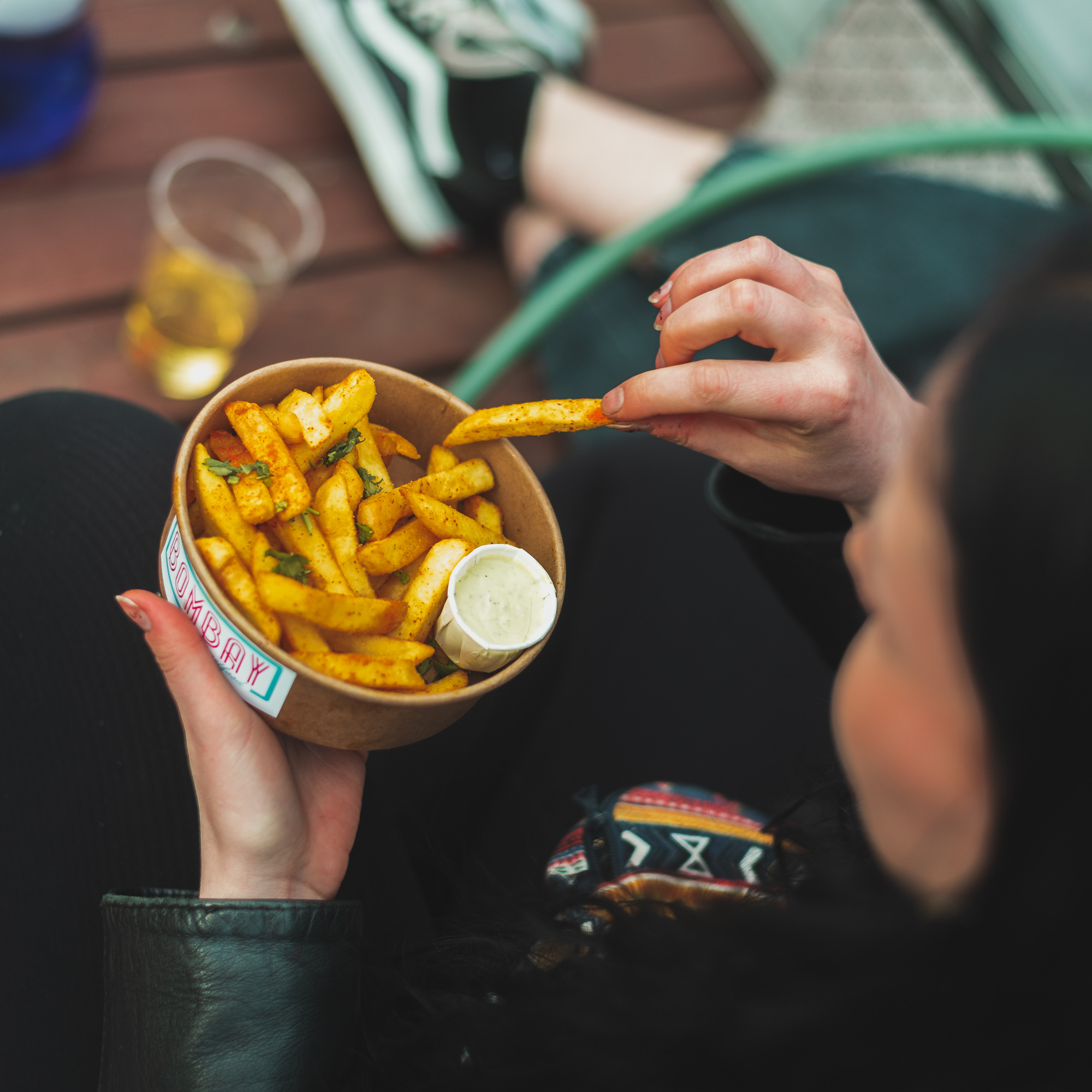 Osoba je frytki z sosem z papierowej miski z logo 'Bombay'. Widok z góry, na drewnianym tle widać napój i fragment butów.