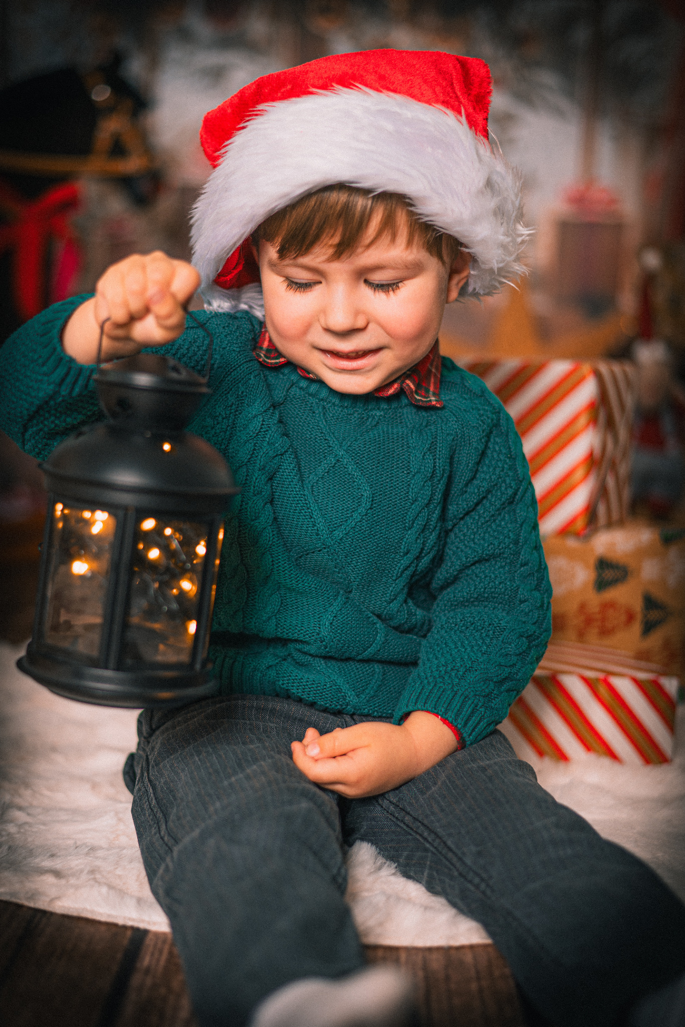 Uśmiechnięty chłopiec w czapce Mikołaja trzyma lampion ze światełkami na tle prezentów. Ubrany w zielony sweter i szarą koszulę w kratę. Klimat świąteczny.