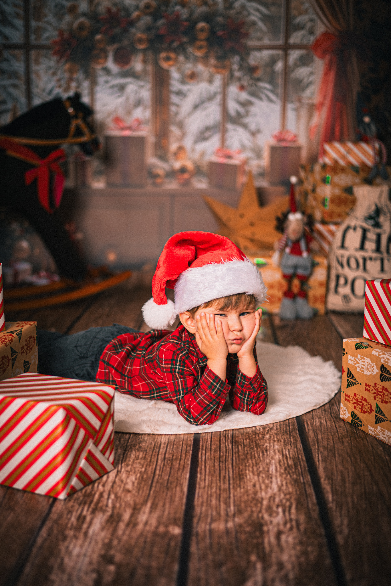 Chłopiec w czapce Mikołaja leży na dywanie wśród prezentów, zamyślony, w świątecznej scenerii z drewnianym koniem i oknem w tle. Ujęcie z niskiej perspektywy.