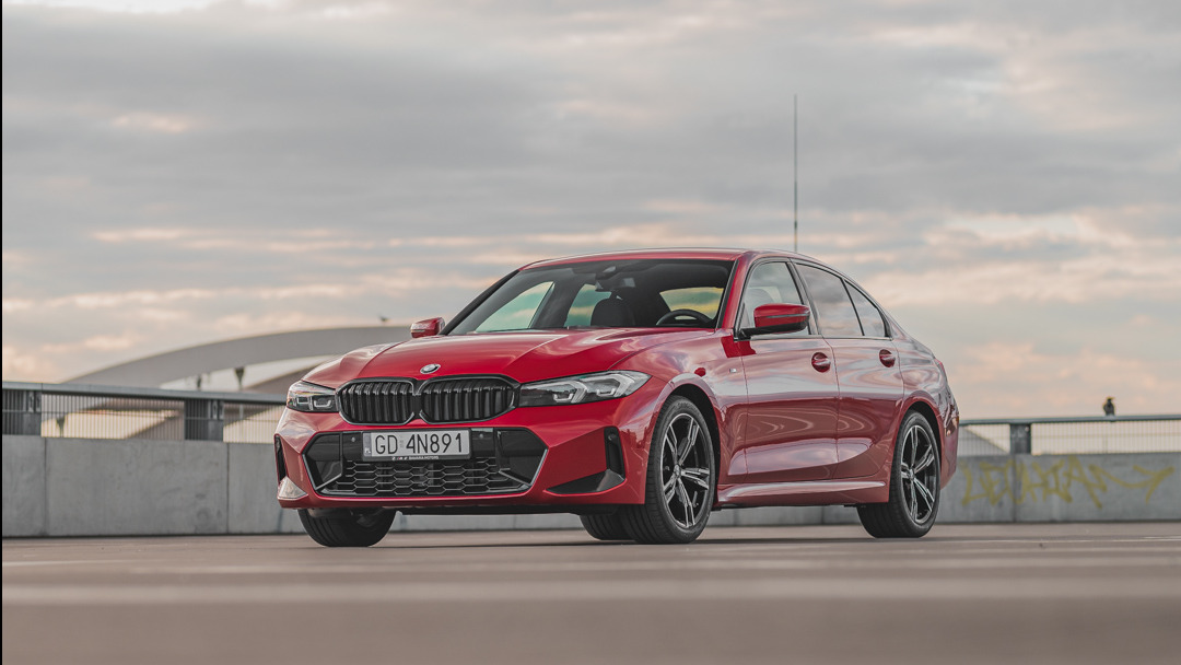 Czerwone BMW serii 3 na parkingu, z widocznym przodem i bokiem auta, na tle pochmurnego nieba i fragmentu mostu. Tablica rejestracyjna widoczna.