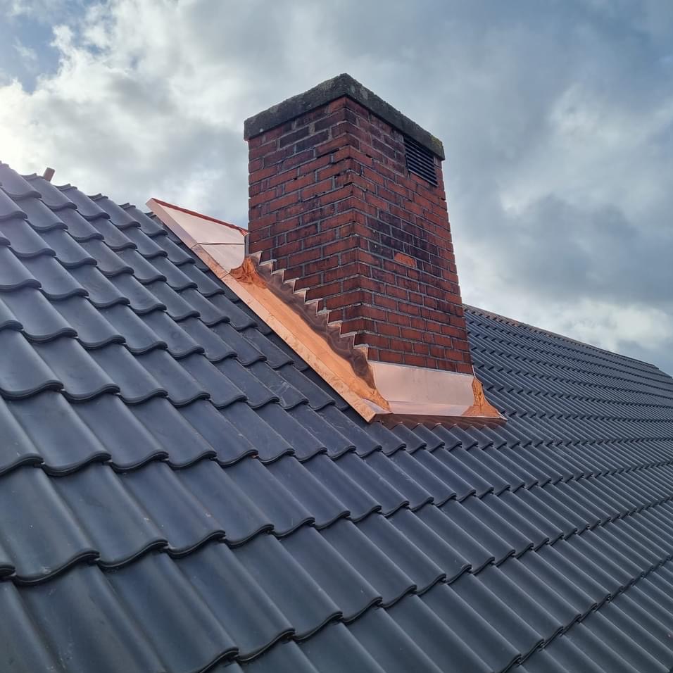 Ciemna dachówka ceramiczna z kominem z czerwonej cegły, obróbka blacharska komina z miedzianymi elementami. Pochmurne niebo w tle.