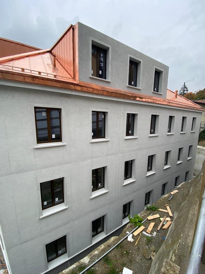 Odnowiona kamienica z nową, miedzianą blachą na dachu i świeżo pomalowaną elewacją w odcieniach szarości. Widoczne nowe okna z ciemnymi ramami.