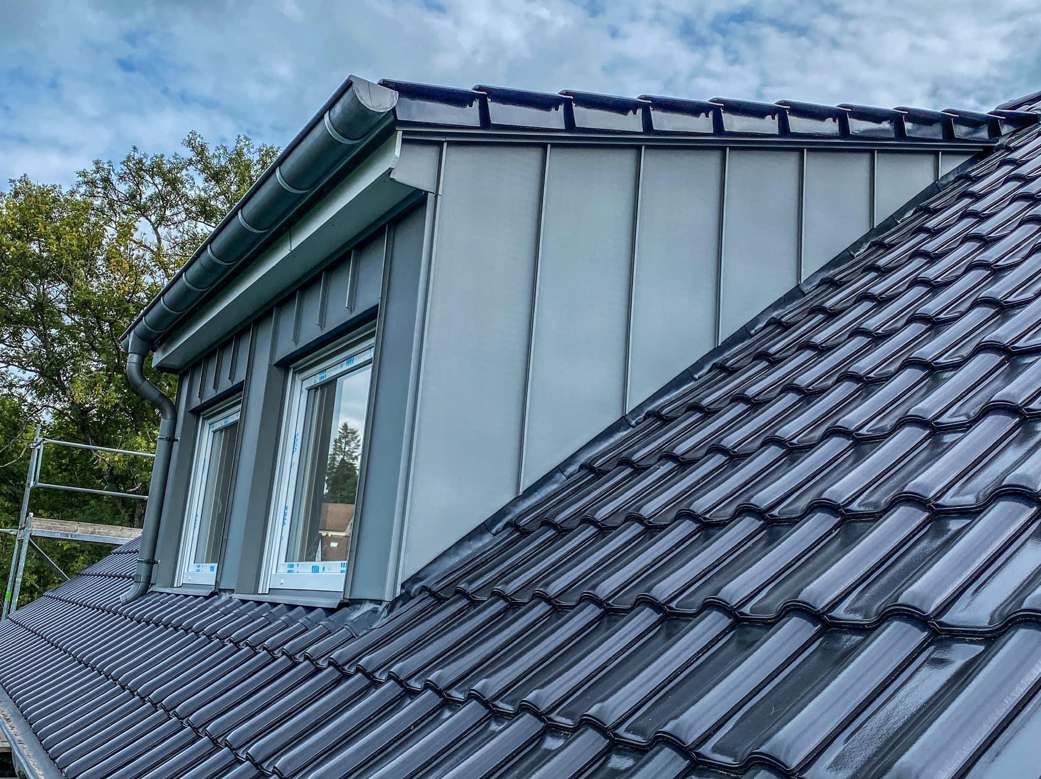 Błyszczący, czarny dach z dachówki ceramicznej sąsiaduje z szarym, metalowym dachem lukarny z oknami. Widoczne rynny i fragment rusztowania.