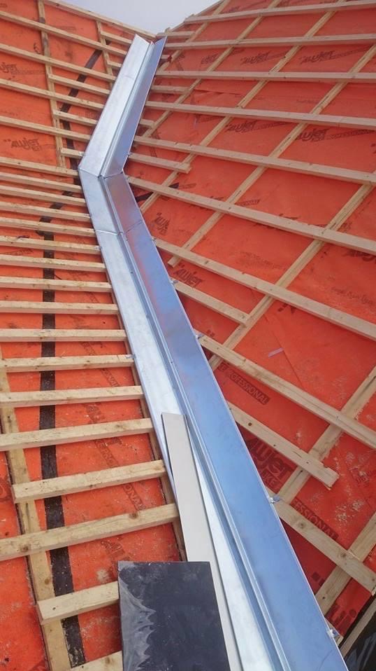 Montaż obróbki blacharskiej na dachu z drewnianą konstrukcją i membraną w kolorze pomarańczowym. Obróbka wykonana z blachy w kolorze srebrnym.