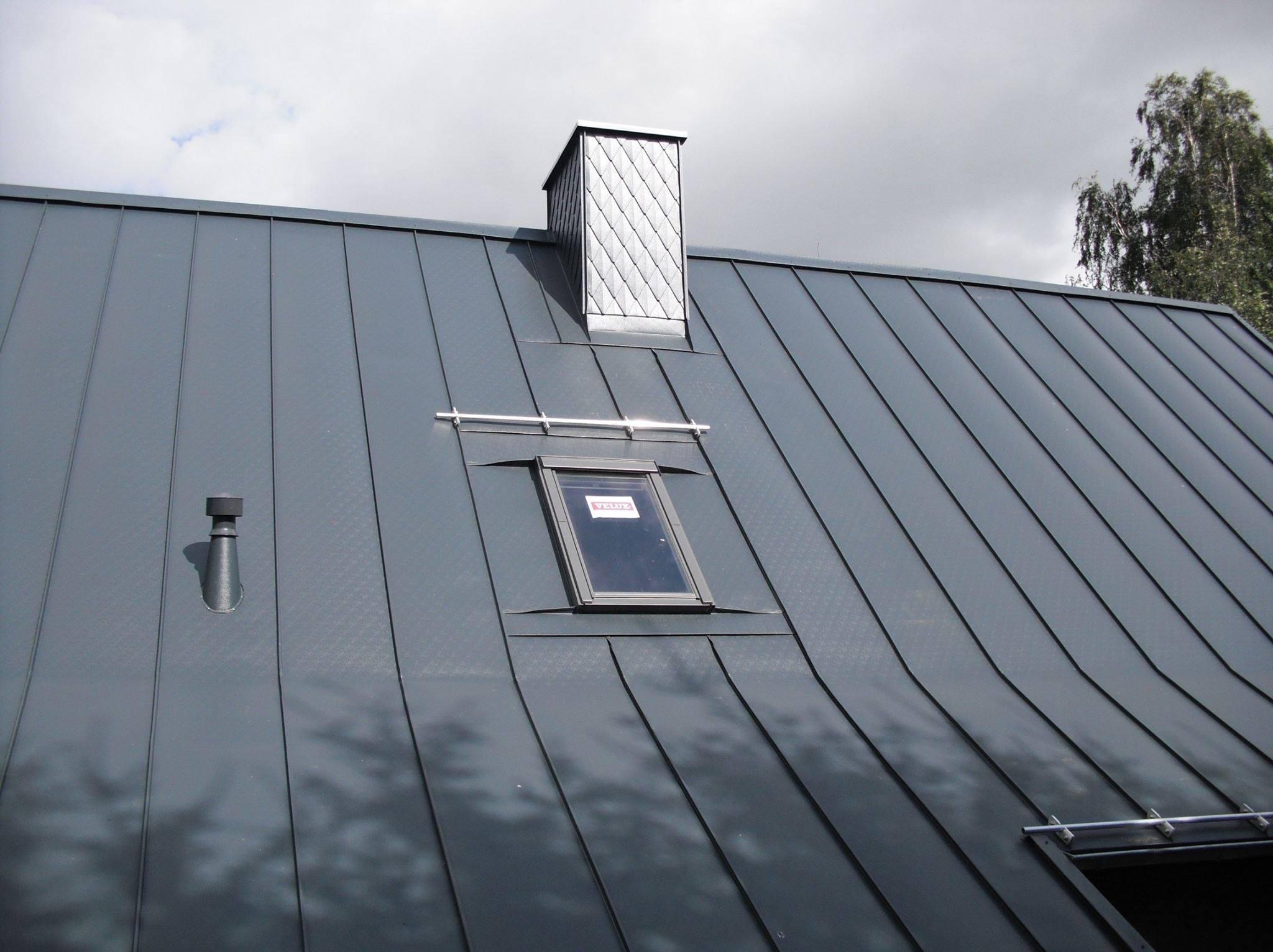 Szary dach z blachy trapezowej z oknem dachowym Velux i kominem obłożonym romboidalną blachą. Widoczne mocowania okna i odpowietrznik. Pochmurne niebo w tle.