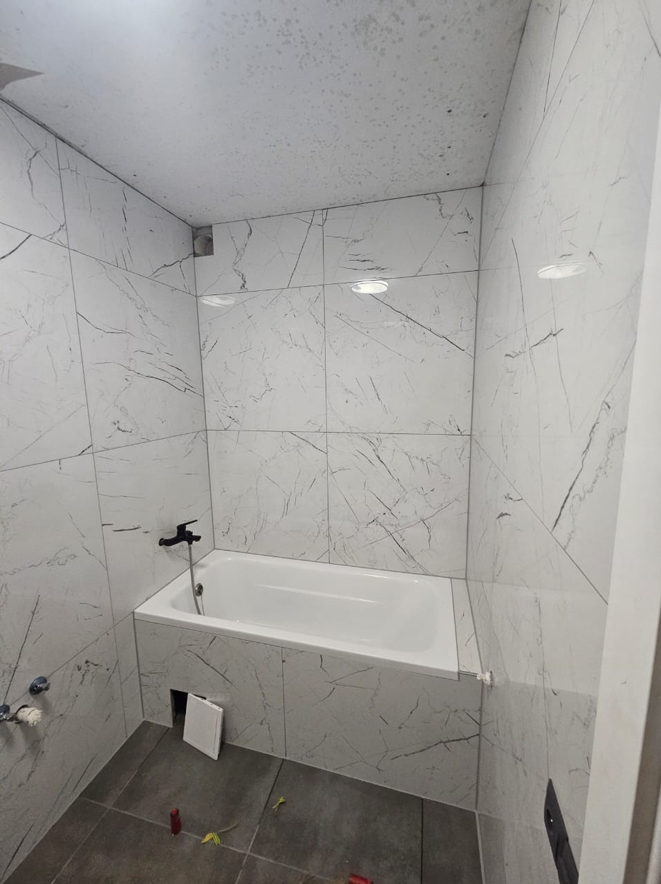 Wykończona łazienka z wanną obudowaną płytkami imitującymi marmur. Szare płytki podłogowe, czarna bateria wannowa. Widoczne detale wykończeniowe i oświetlenie.