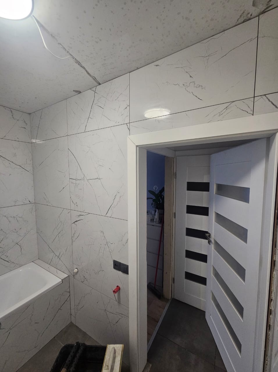 Wykończona łazienka z wanną i białymi płytkami imitującymi marmur na ścianach i suficie. Widoczne otwarte białe drzwi z poziomymi przeszkleniami.