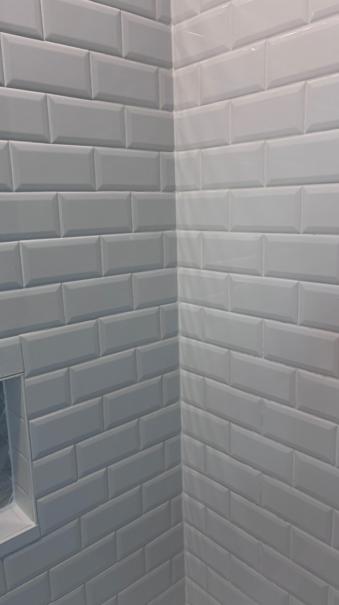 Białe, błyszczące płytki typu metro ułożone w narożniku łazienki, z widoczną wnęką wykończoną tym samym materiałem. Minimalistyczny design.