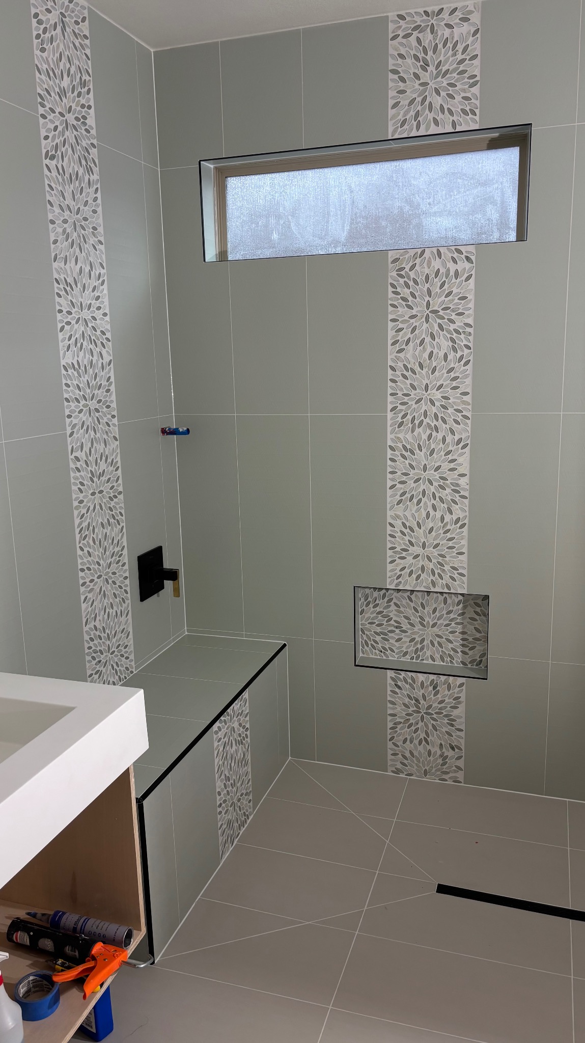 Wykończona łazienka z szarymi płytkami i dekoracyjnymi pasami mozaiki. Wnęka w ścianie wyłożona mozaiką. Widoczny fragment szafki pod umywalkę i okno.