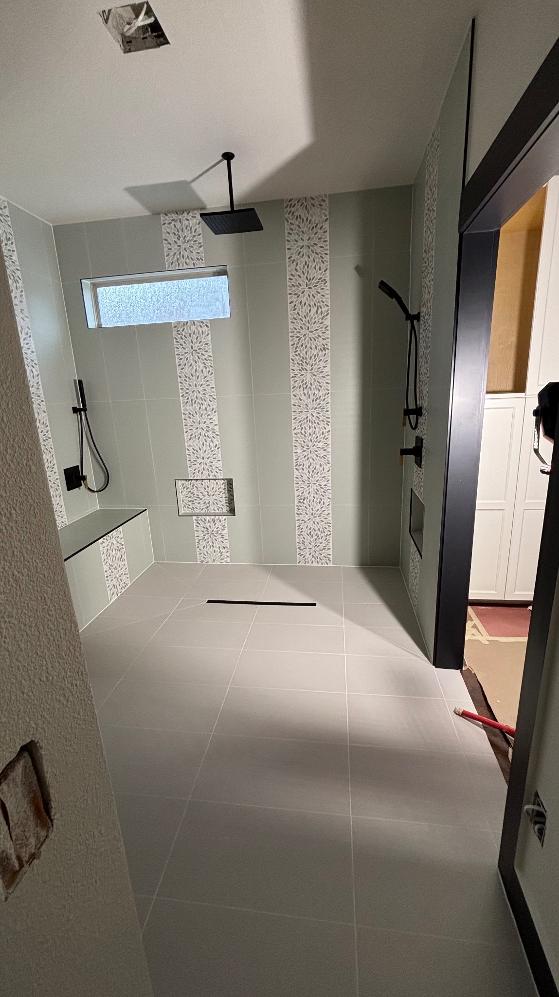 Nowoczesna łazienka z szarymi płytkami, czarną armaturą i dekoracyjnymi pasami płytek. Widoczny odpływ liniowy i wnęka w ścianie. Minimalistyczny design.