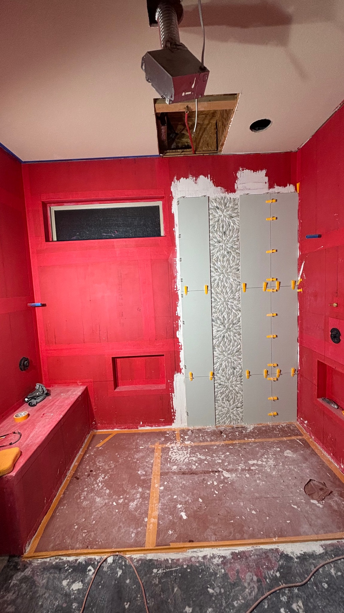 Remont łazienki w trakcie: czerwona hydroizolacja, układanie szarych płytek z wzorem, żółte krzyżyki dystansowe, widoczne narzędzia i instalacja. Otwór w suficie.