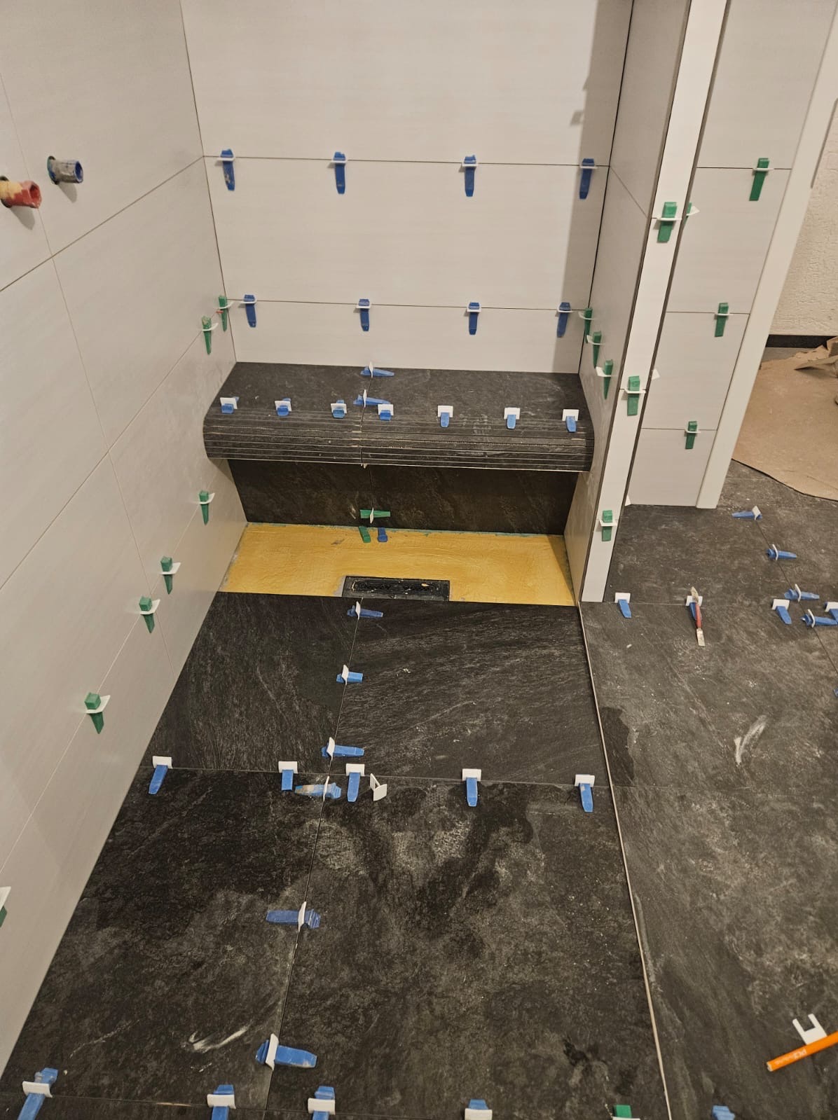 Układanie ciemnych płytek w łazience z półką w Łasku. Widoczne klipsy poziomujące i odpływ liniowy. Białe płytki na ścianach, etap prac.